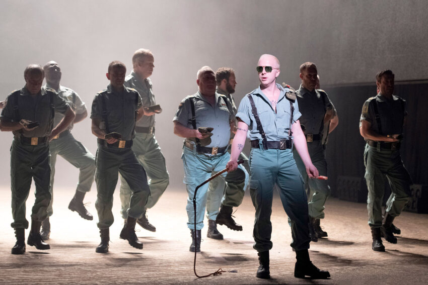 Male performer dressed in an army uniform holding a whip with other male performers behind